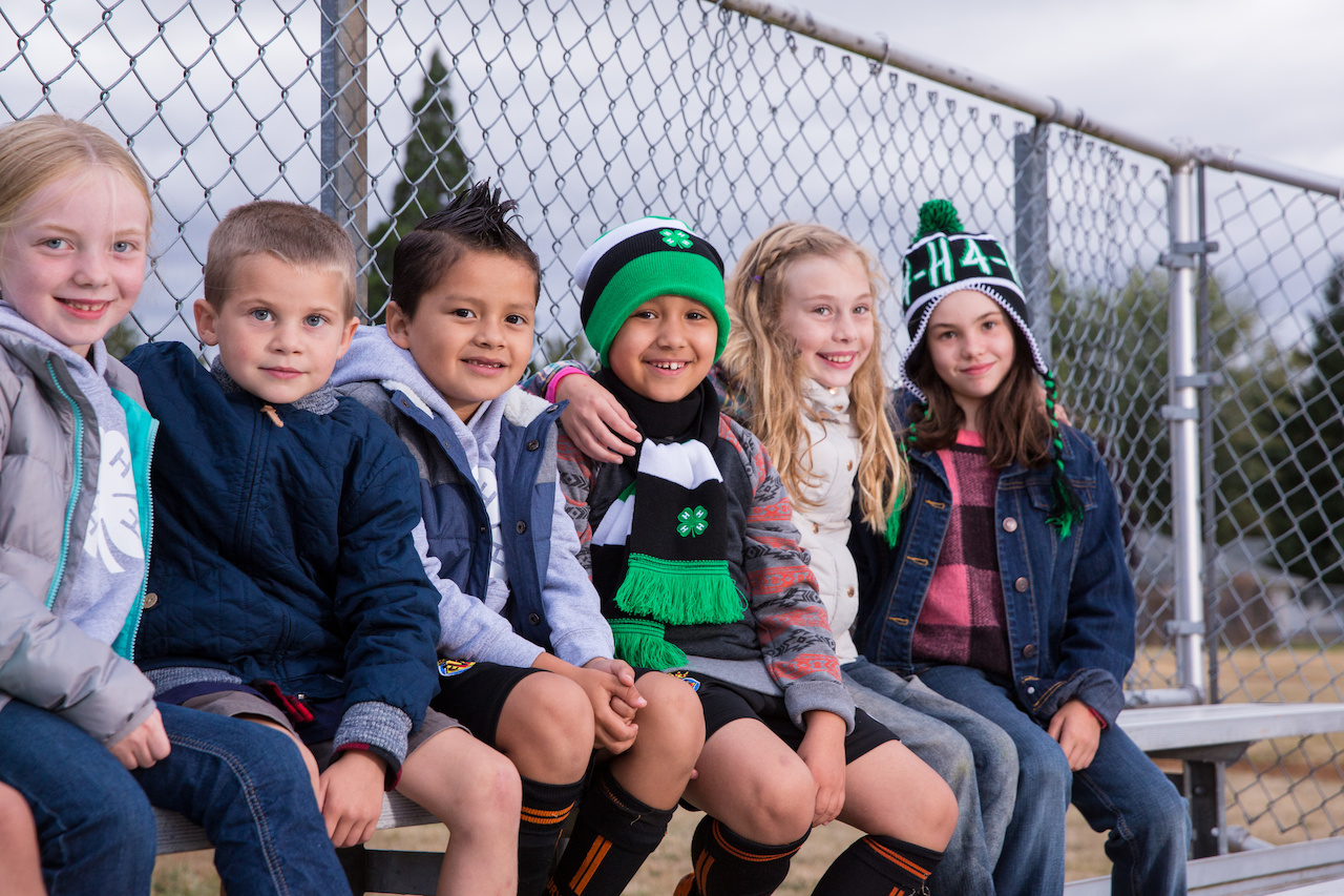 Six kids sit together on a bench at a sports field